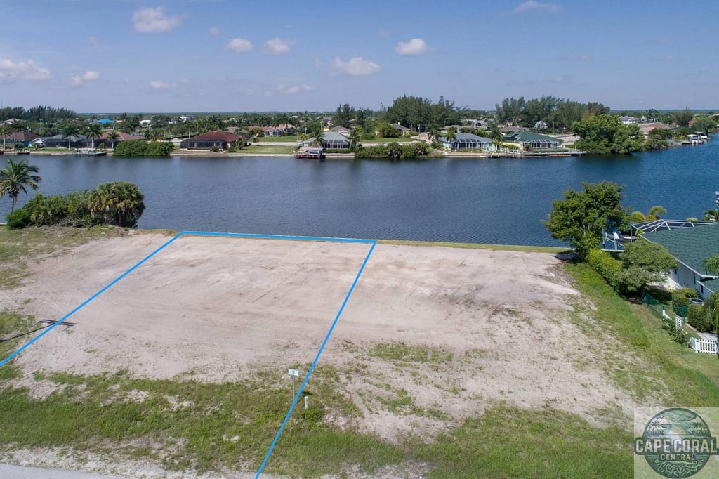 Aerial view of a vacant residential lot highlighted in blue, available for sale in North Cape Coral, surrounded by lush greenery and established roadways.