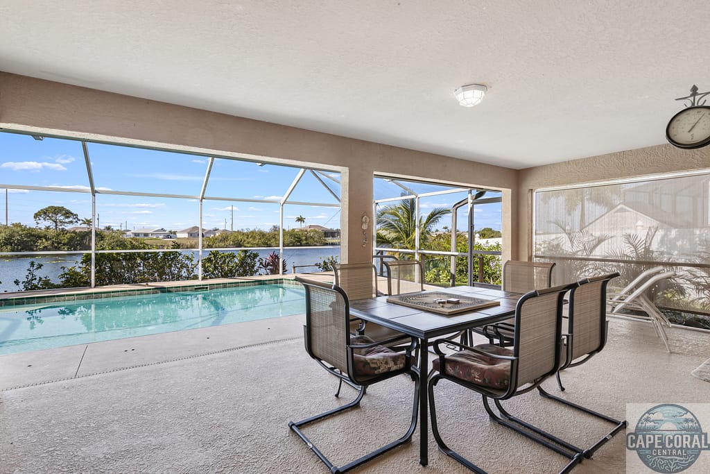 Screened-in dining area with a table set by a pool overlooking a calm canal in a residential Cape Coral neighborhood, under a clear sky