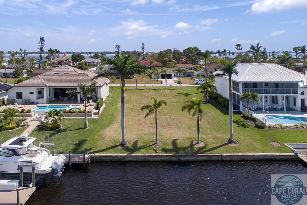 Elevated view of a direct access waterfront lot for sale in Cape Coral, FL, showcasing the canal frontage, adjacent upscale homes, and ready-to-build land with lush greenery.