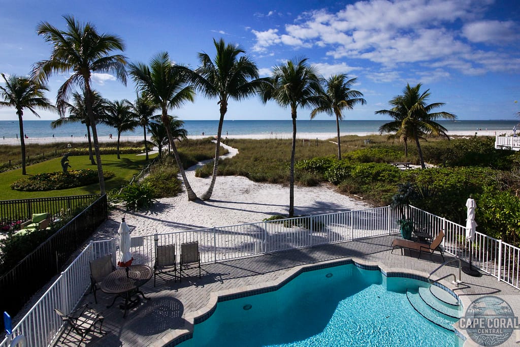 Fort Myers beachfront home for sale with a pool, patio furniture, and direct beach access, flanked by lush greenery and tall palm trees with a clear view of the Gulf waters
