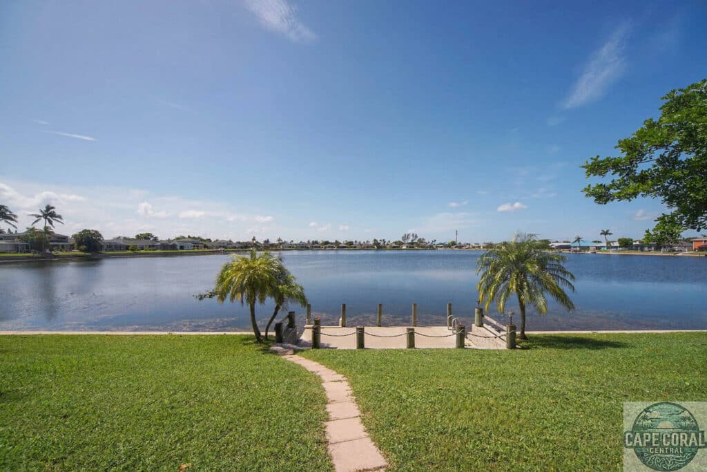 Lush green lawn leading to a private dock on a tranquil lake, flanked by palm trees, with residential homes in the distance off Trafalgar