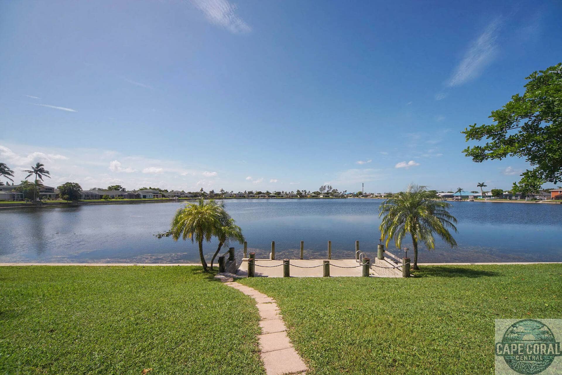 Lush green lawn leading to a private dock on a tranquil lake, flanked by palm trees, with residential homes in the distance off Trafalgar